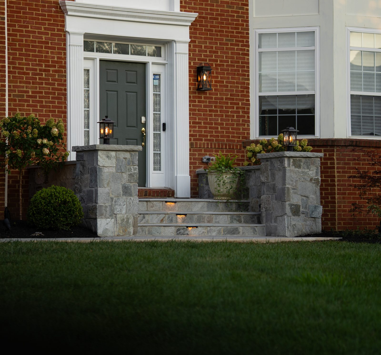 Stone entry steps and lit plantings at a brick home at dusk